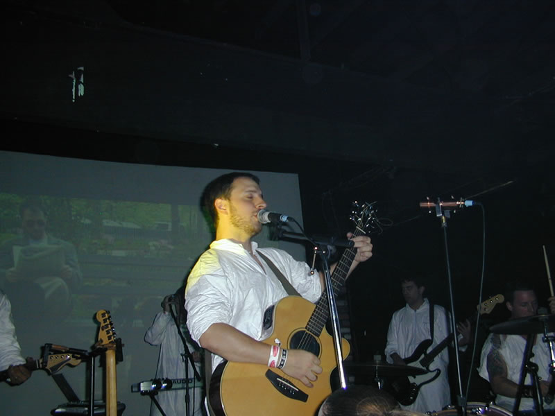 Burns singing whilst playing guitar. There are images projected behind him.
