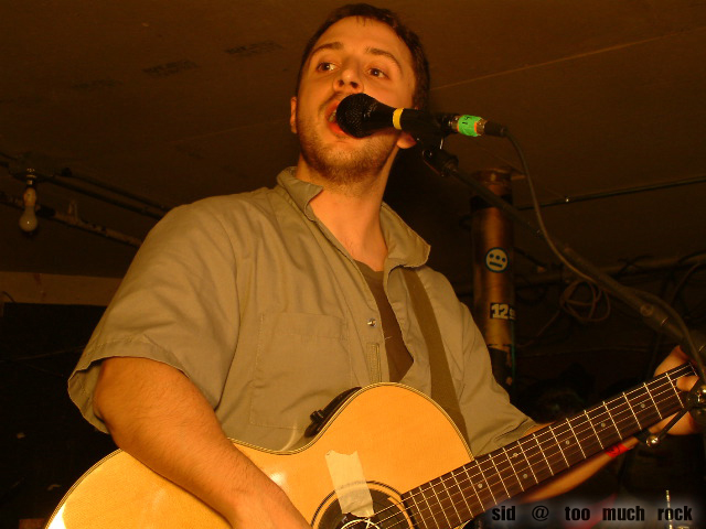 Burns playing acoustic guitar and singing into the microphone on stage.