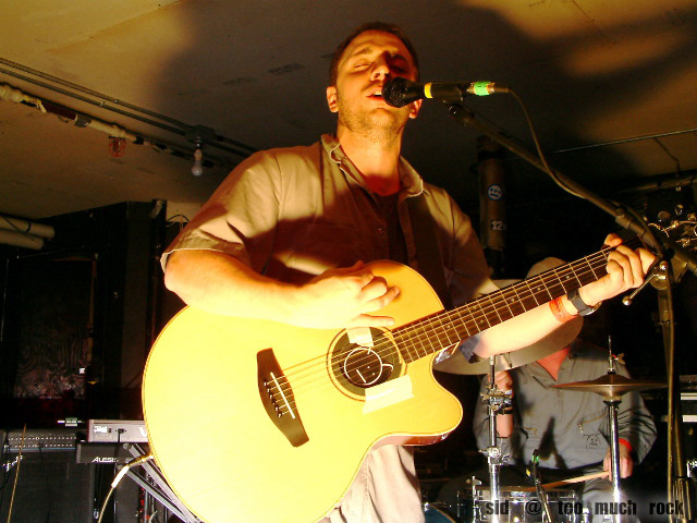 Burns playing acoustic guitar and singing into the microphone on stage.