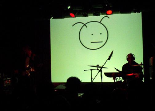 Burns with band on stage. An image of a dustmite is projected on the screen behind them.