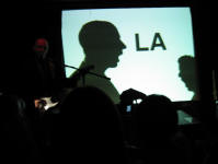 Burns on stage whilst the image of two silhouettes saying LA is projected on the screen behind him.