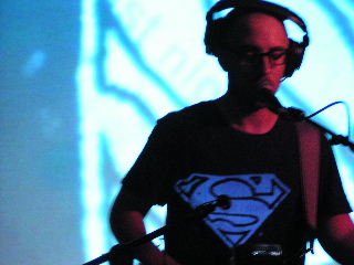 Burns on stage. He has the Superman logo projected on his shirt.