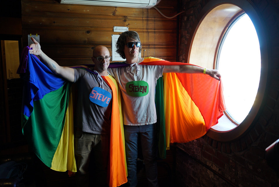 Steve Burns and Steven Drozd standing close together. They are both wearing a rainbow cape and each have one arm stretched out to expand the cape. They also have large name tags connected to their chests.