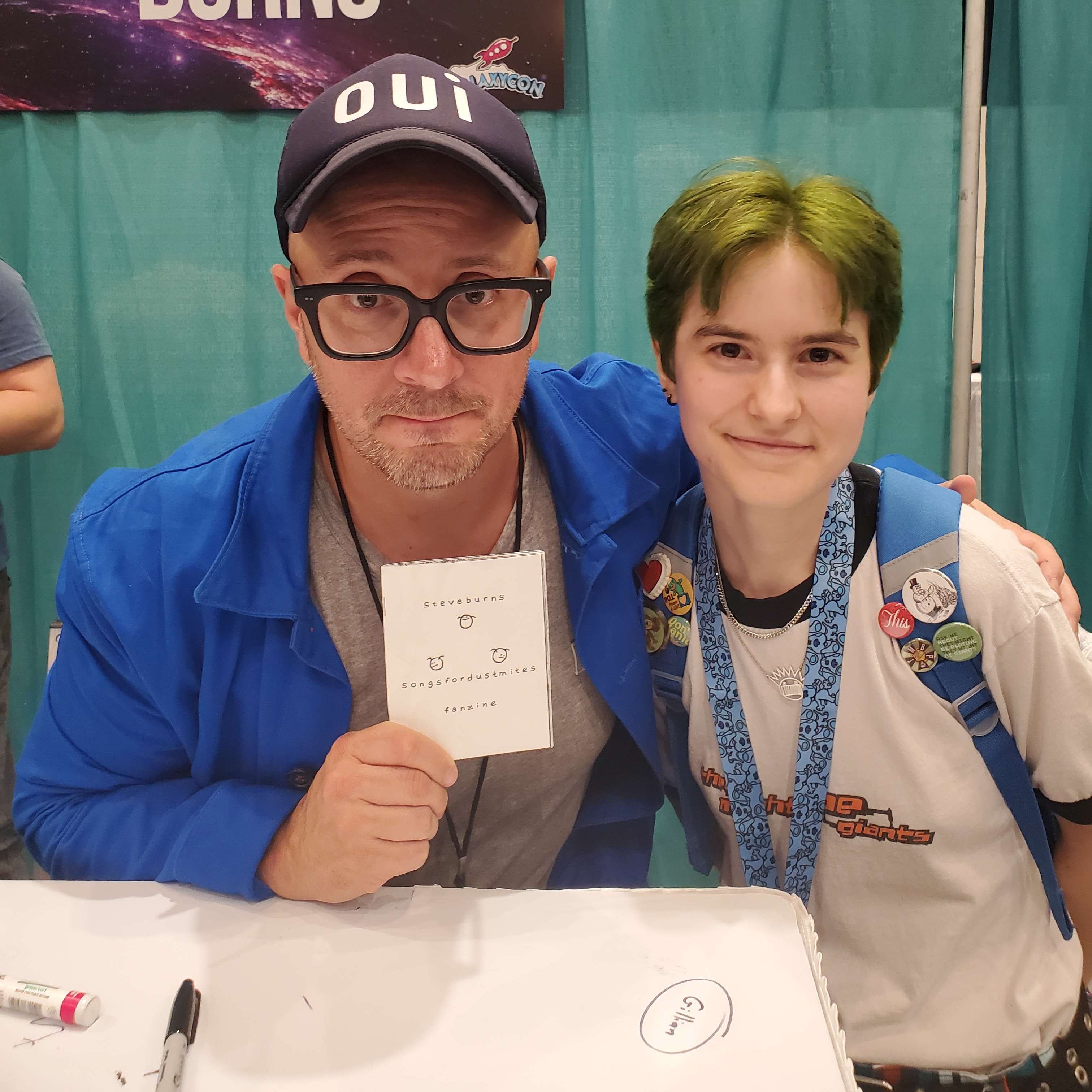 Steve Burns and Seymour posing together and smiling for the camera with Steve on the left and Seymour on the right. Steve is holding up a tiny zine in front of him and has his arm wrapped around Seymour.