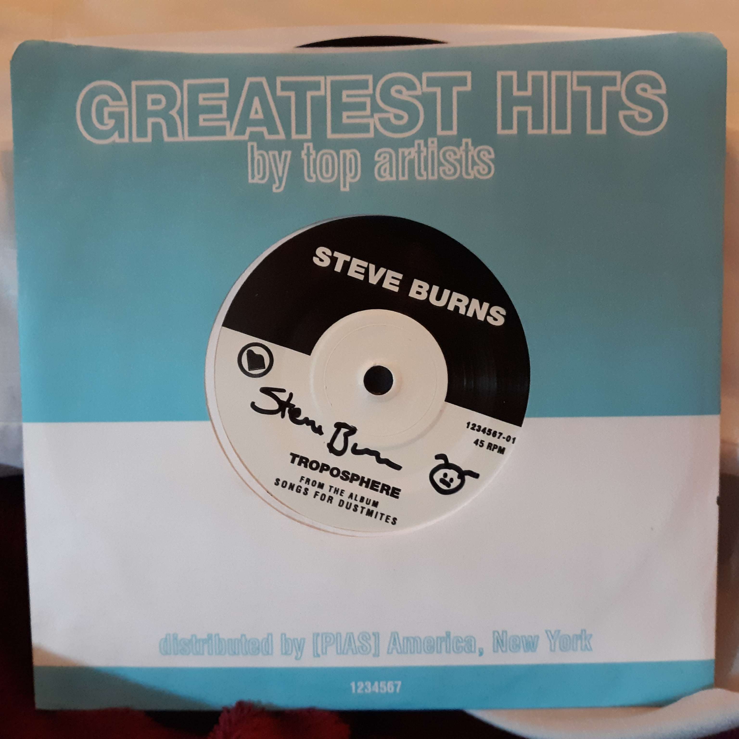A seven inch single within its paper slip. The paper slip says 'Greatest Hits by top artists' on the top with 'distributed by PIAS America, New York' on the bottom. The label on the single itself says 'Steve Burns' on the top with 'Troposphere from the album Songs For Dustmites' on the bottom. Steve Burns' signature is in between them with a drawn dustmite next to his name.