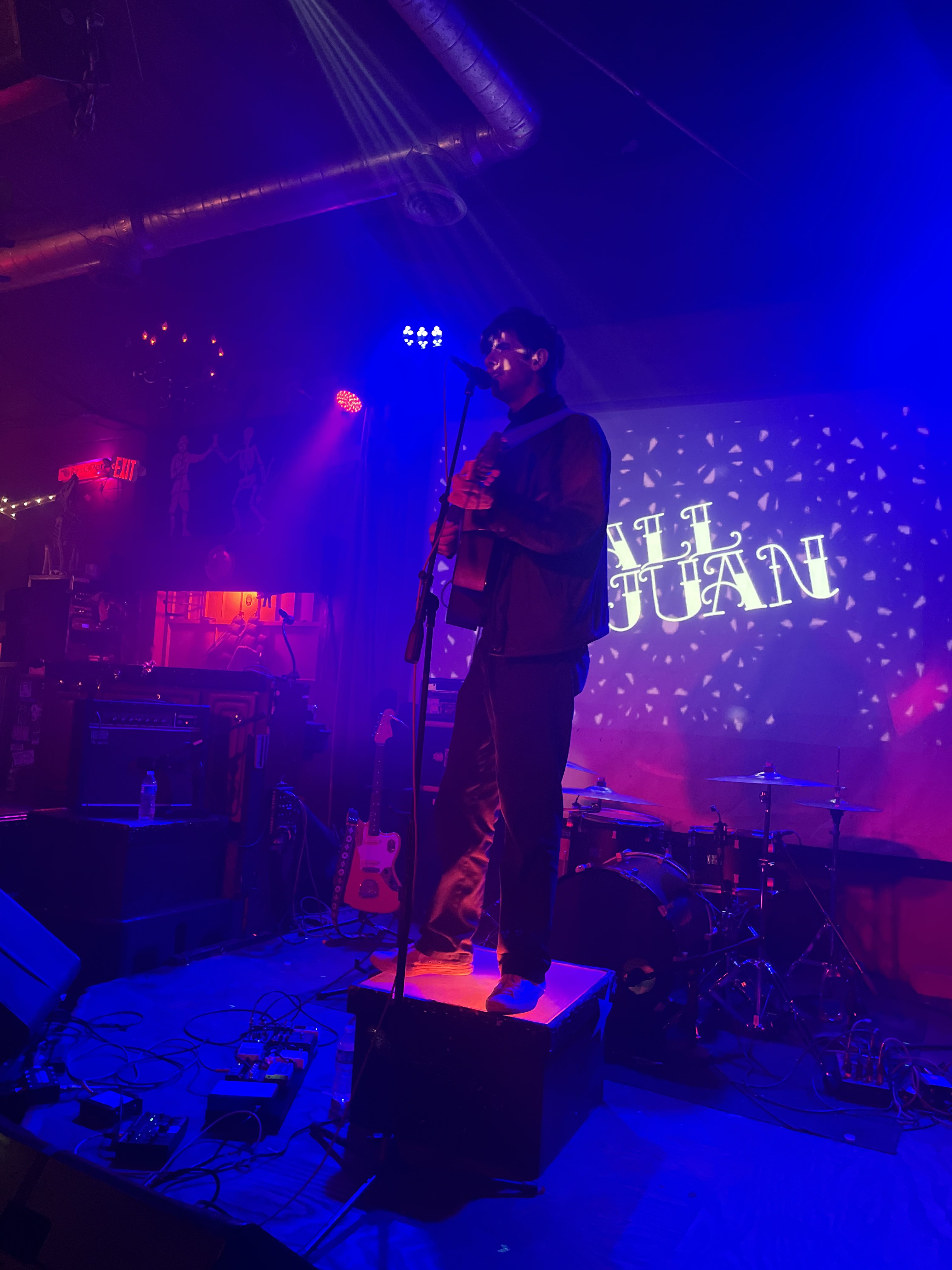 Tall Juan on stage playing a guitar and singing into a microphone. His name is projected on the wall behind him.