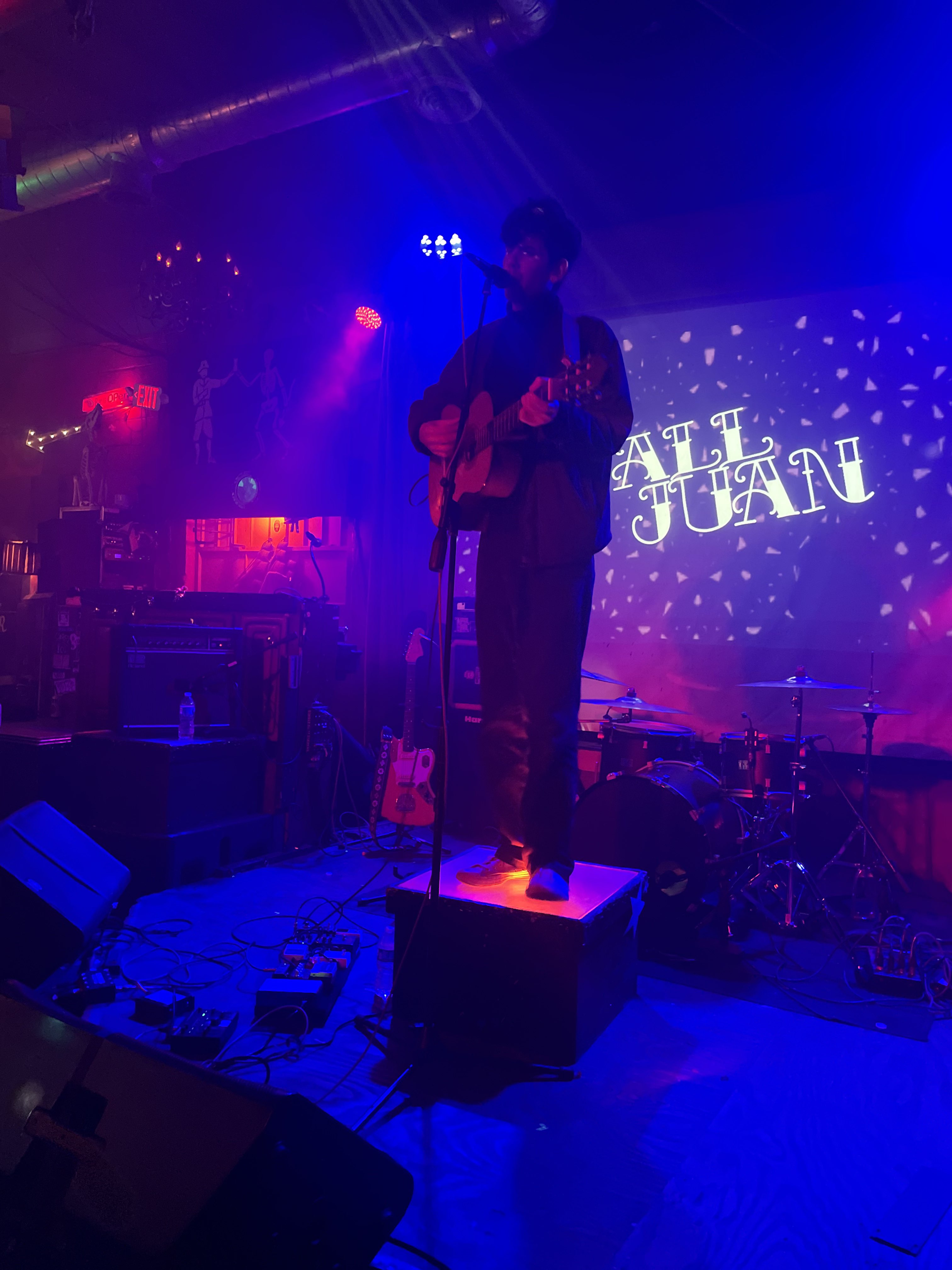 Tall Juan on stage playing a guitar and singing into a microphone. His name is projected on the wall behind him.