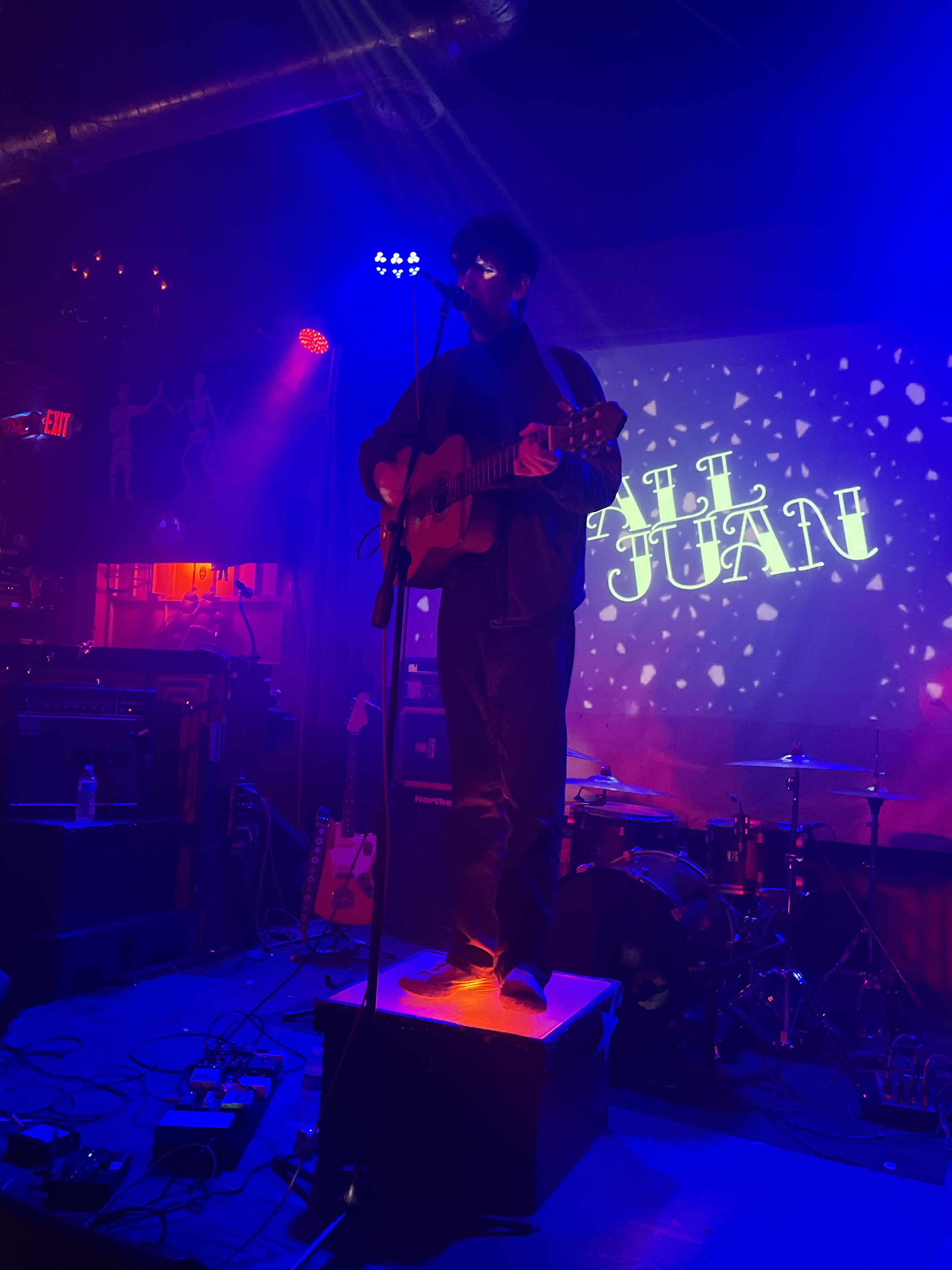 Tall Juan on stage playing a guitar and singing into a microphone. His name is projected on the wall behind him.