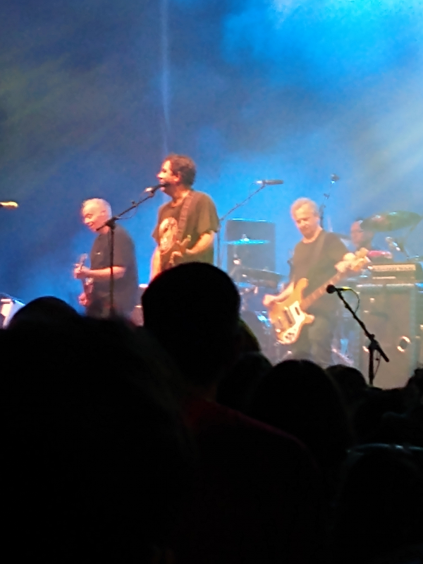 Ween on stage. Gene Ween is on the left, Dean Ween is in the middle, and Dave Dreiwitz is on the right. Claude Coleman is in the back on the drums. The bottom half of the photo is just the dark heads of people in the crowd.