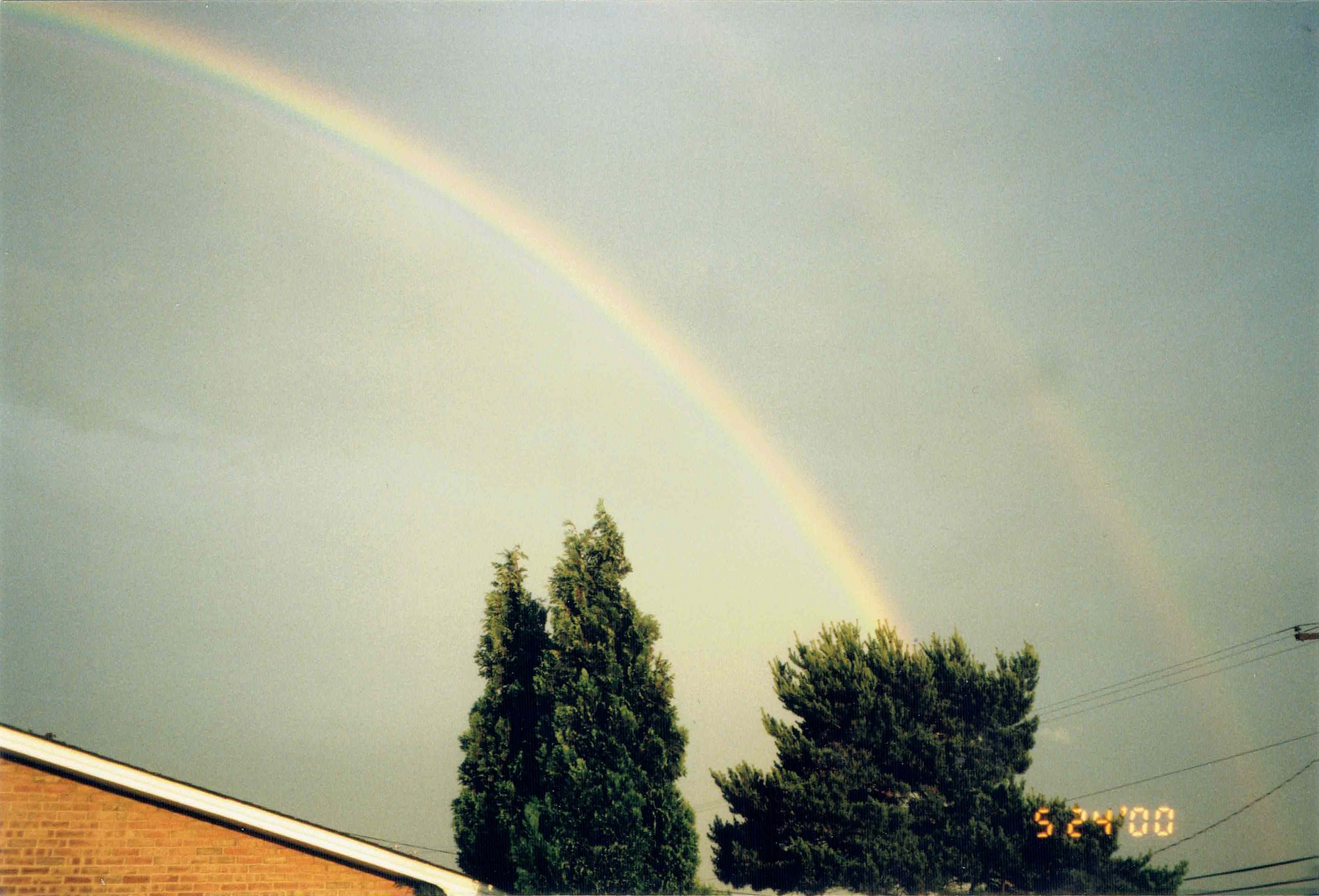 A double rainbow in the sky. There are some trees and the roof of a house below them.