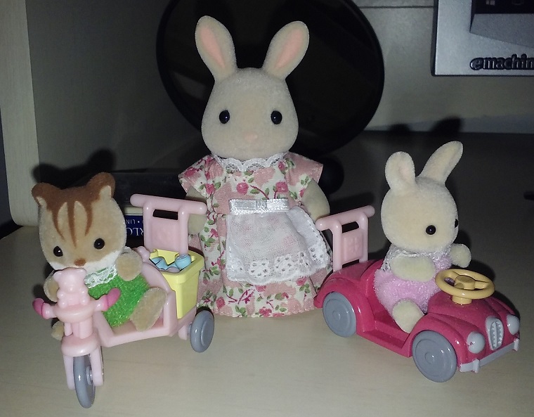 Three Calico Critters on a desk. Jake the squirrel is on the left in his pink tricycle that has a yellow basket in the back. Jake is wearing a green outfit. Apple the bunny is on the right in her dark pink car with yellow steering wheel. She is wearing a pink outfit. Gwyneth the bunny is behind them in the middle. She is the mother of Apple and is wearing a pink floral dress with white apron.