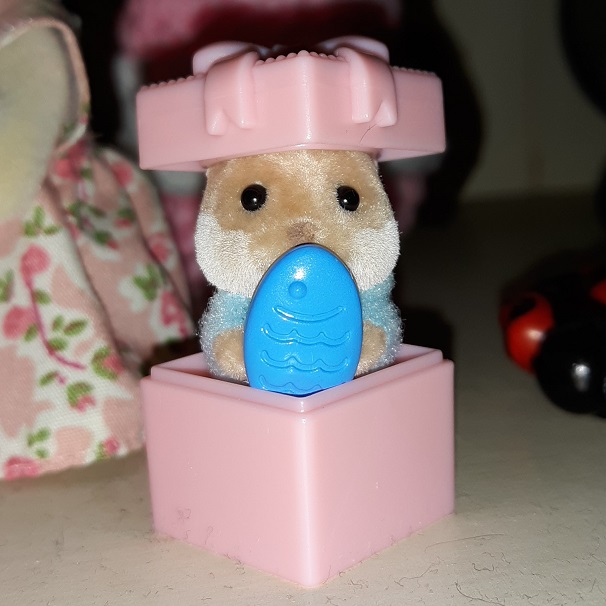 An otter in a pink gift box. The top of the gift box is sitting on their head. They are holding a blue fish in front of them. They are wearing a blue outfit.