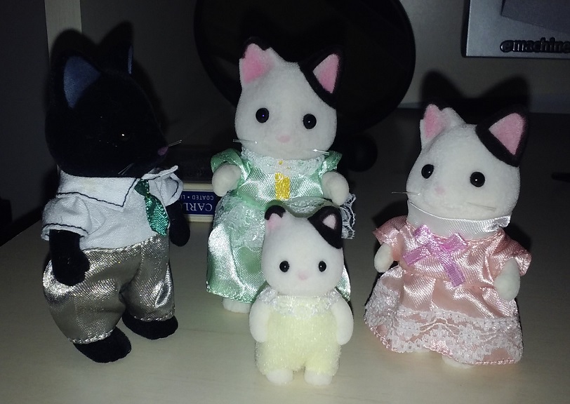 A family of black and white cats. The father is on the far left and is all black. He is wearing a short sleeve white shirt with a green tie. He is wearing silver pants. The mother is right next to him. She is white with black spots. She is wearing a green dress. The older daughter is on the right and looks the same as the mother, but is slightly shorter and is wearing a pink dress. The youngest daughter is in the front. She also looks like the mother. She is wearing a yellow outfit.