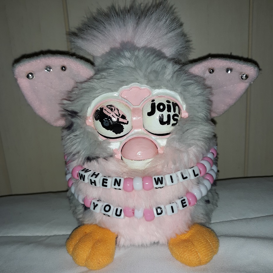 A grey and pink furby with a painted pink and white faceplate. They have a monster truck hearse painted on one eyelid and the words 'join us' painted on the other. They have two pink and white beaded necklaces that says 'when will you die.'