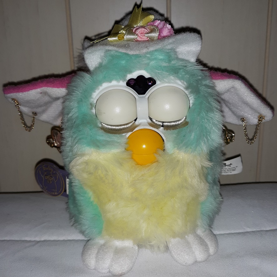 A pale green and yellow furby with bright pink ears. They are wearing a white hat with flowers and a yellow bow on it.