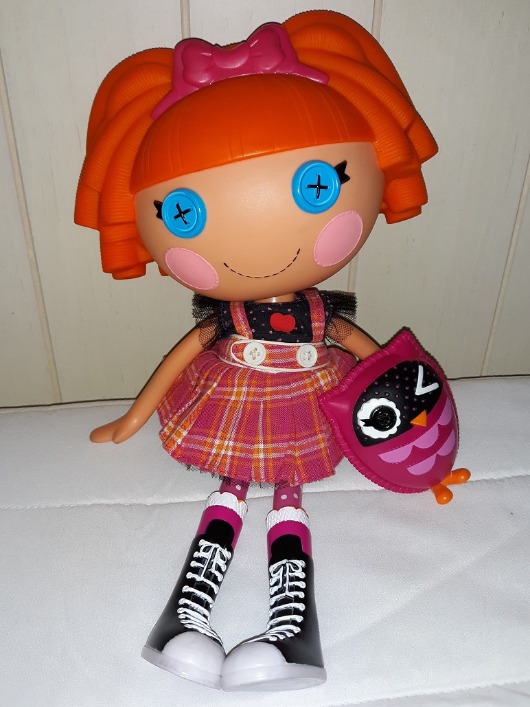 Bea sitting on a bed with her pink owl. She is light skinned with orange hair that has a straiht blunt fringe and is curly in the back and has a pink bow in her hair. She has blue button eyes. She is wearing a black shirt with an apple graphic on the chest and is wearing a pink and orange tartan overall skirt. She has black lace up sneakers that go up to her knees.
