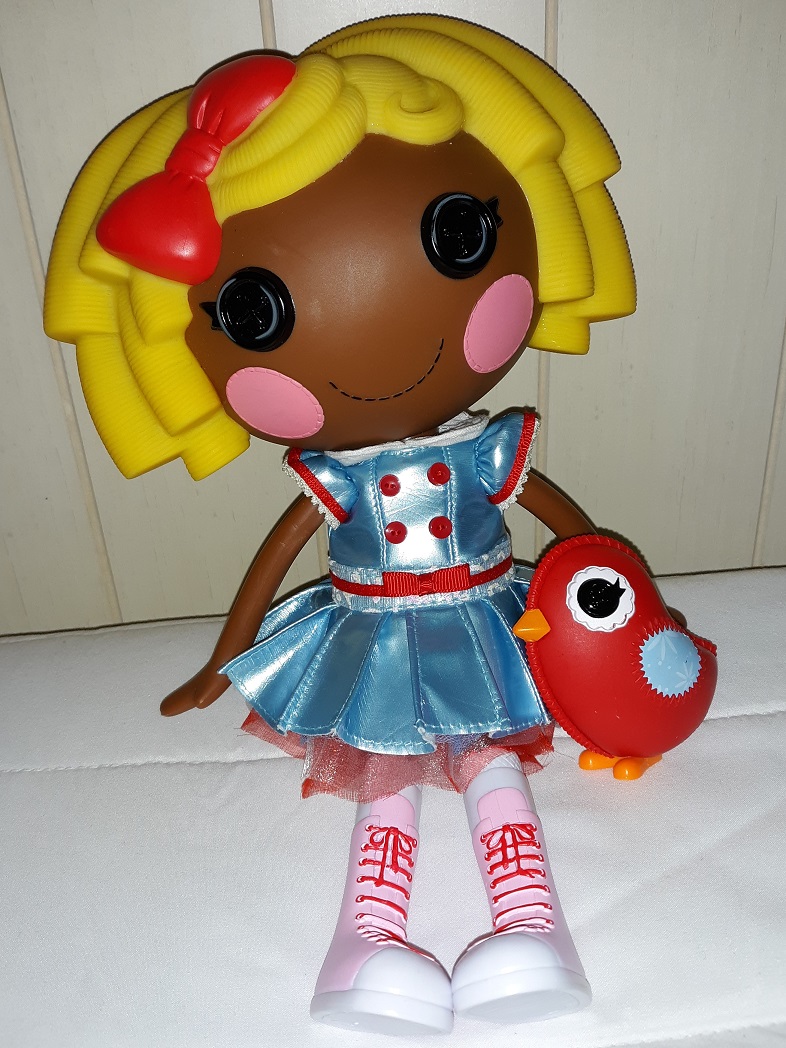 Dot sitting on a bed with her red bird. She is dark skinned with yellow hair and has a large red bow in it. She has black button eyes. She is wearing a light blue chromatic dress with red buttons on the chest, a red belt on the waist, red trim on the sleeves, and red tulle underneath. She has on pink lace up sneakers that go up to her knees.