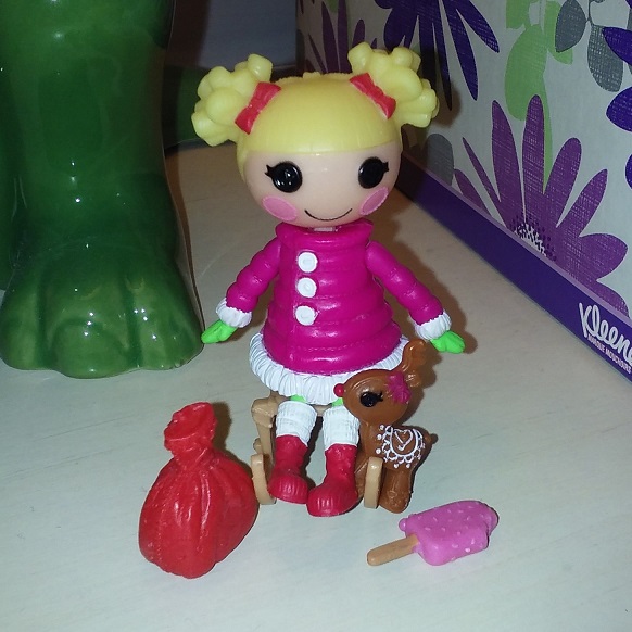 Holly s light skinned with blonde hair. She has a straight blunt fringe with two puffs of curly hair tied up with pink bows. She is wearing a large pink puffy coat that has white buttons and white trim on the sleeves and at the bottom of the coat. She has green gloves and green tights. She is wearing red boots. She is sitting on a sleigh and has a reindeer next to her with a red sack and pink icepop.