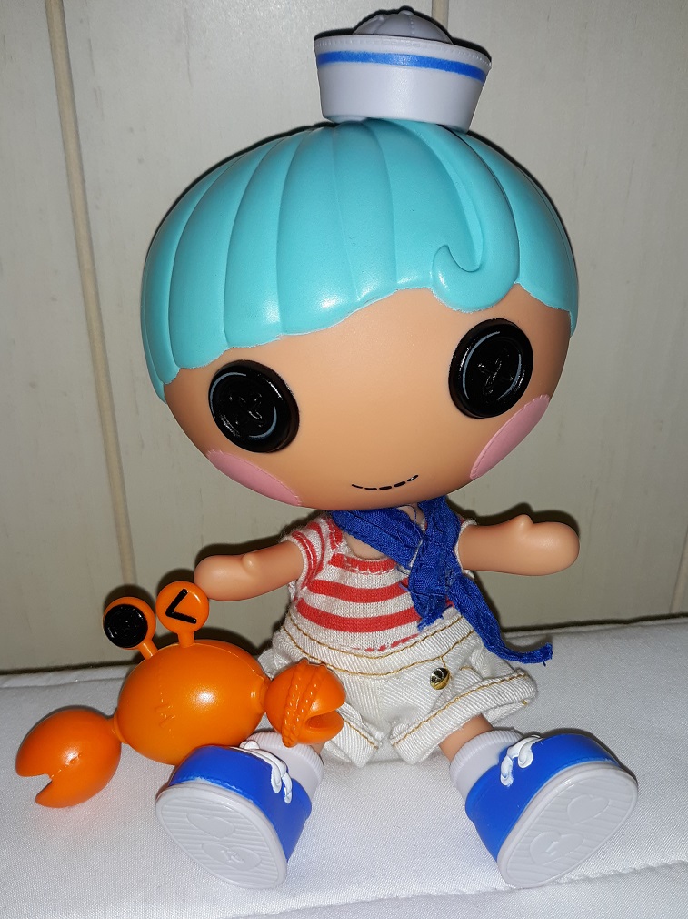 Matey sitting on a bed with his orange crab. He is light skinned with light blue hair and a sailor hat. He has black button eyes. He is wearing a blue ascot, a red and white striped shirt, and white shorts. He has on blue sneakers with white socks.