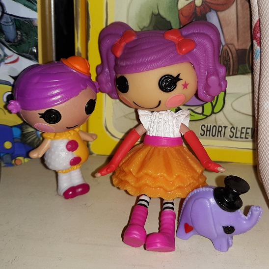 Two Lalaloopsy toys with Squirt Lil Top on the left and Peanut Big Top on the right with their elephant. Squirt has purple hair and is wearing an orange cap. She is wearing a yellow and white outfit with pink buttons. She is wearing white tights and pink shoes. Peanut also has purple hair and is wearing two red bows. She has a white top with an orange skirt and a pink belt on. She is wearing two red arm warmers and has black and white striped tights. She also has on pink boots. Their elephant is purple with a black top hat on.