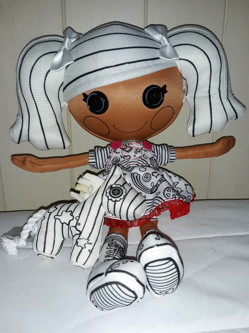 Spot sitting on a bed with her black and white horse. She has a medium skin tone with black button eyes. Her hair is in two pony tails with a bow on each side. Her whole outfit is in black and white besides the red buttons, some stitching, and the red trim at the bottom of her dress. She is wearing a striped shirt with an overall dress that has paintbrushes and palettes on it. She has lace up shoes on that go up to her knees.