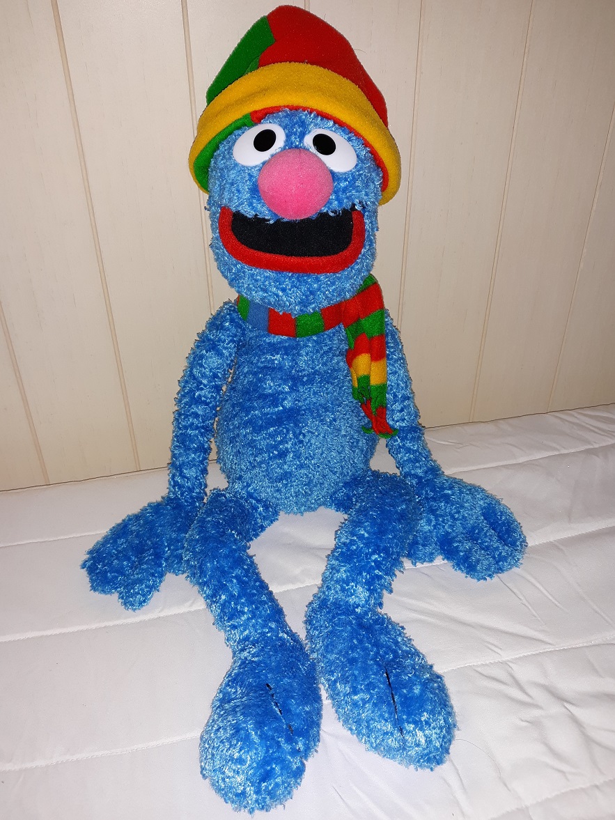 A very large plush doll of Grover that is wearing a green, red, and yellow fleece hat with a matching scarf.