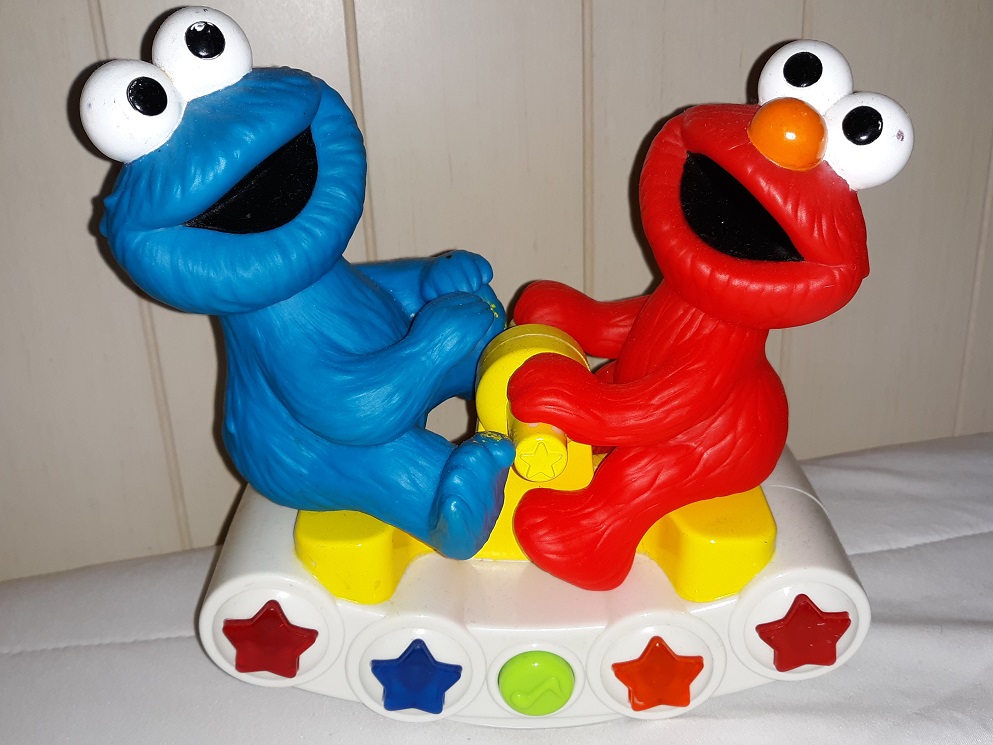 A plastic toy of Cookie Monster and Elmo on a seesaw that has stars and a button on the side of it.