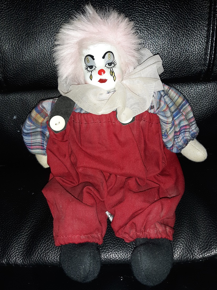 A clown with a porcelain face and fabric body. They have fluffly light pink hair, red overalls, and a striped shirt underneath.