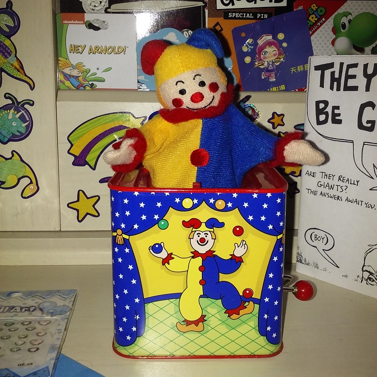 A clown jack-in-the-box that is red, yellow, and blue. The box has a drawing of them on it juggling three balls.