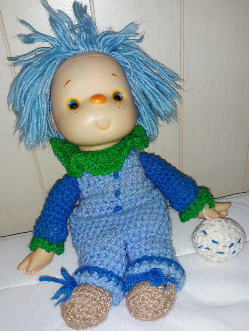 A clown doll with a plastic head and hands with the rest made of yarn. He has blue hair and a blue outfit with green trim. He is holding a white ball.