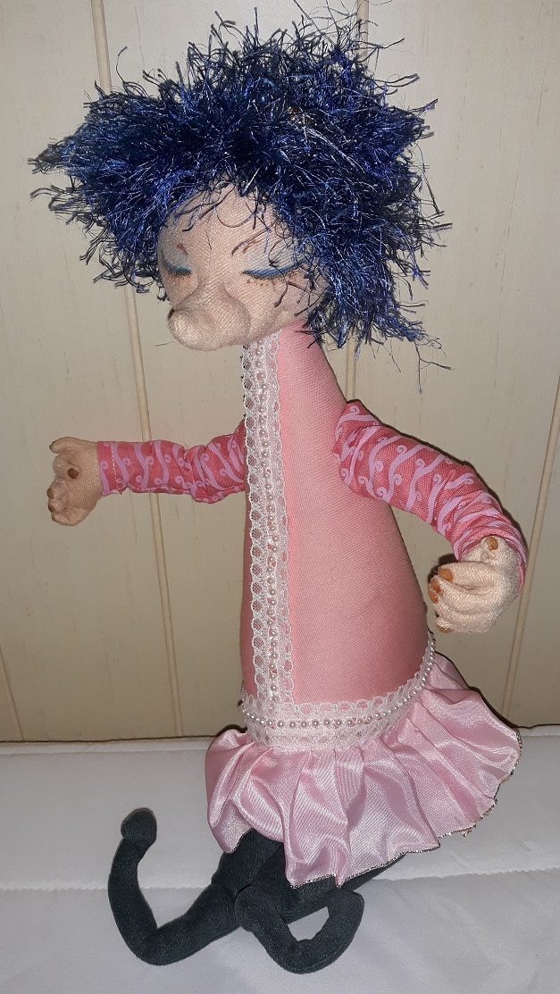 A doll of a woman with a long pointed nose and blue scraggly hair. She is wearing a pink dress with pearls and lace going down the center and around the bottom of it and a pleated pink trim. Her legs and feet are grey.