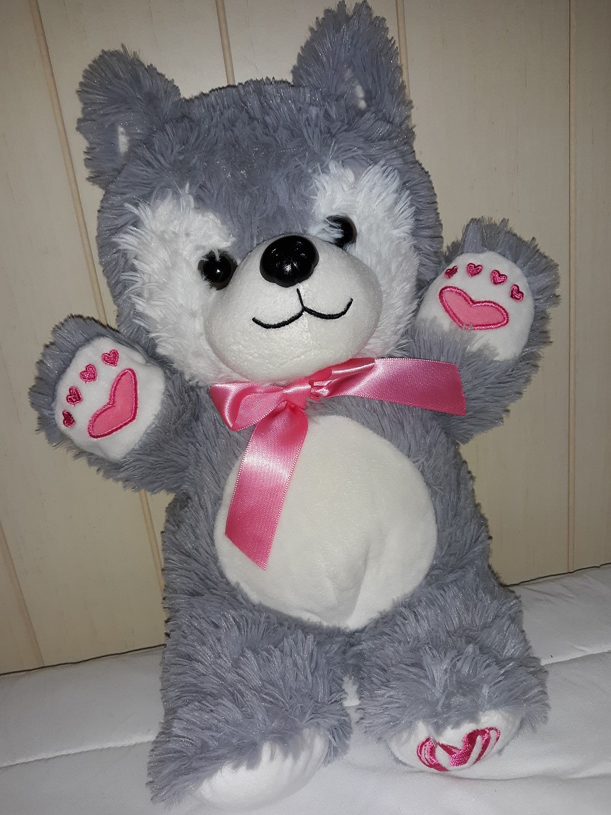 A large doll of a cutesy grey and white wolf. She is wearing a pink bow. Her short arms are up and you can see that her toe pads are all heart shaped.
